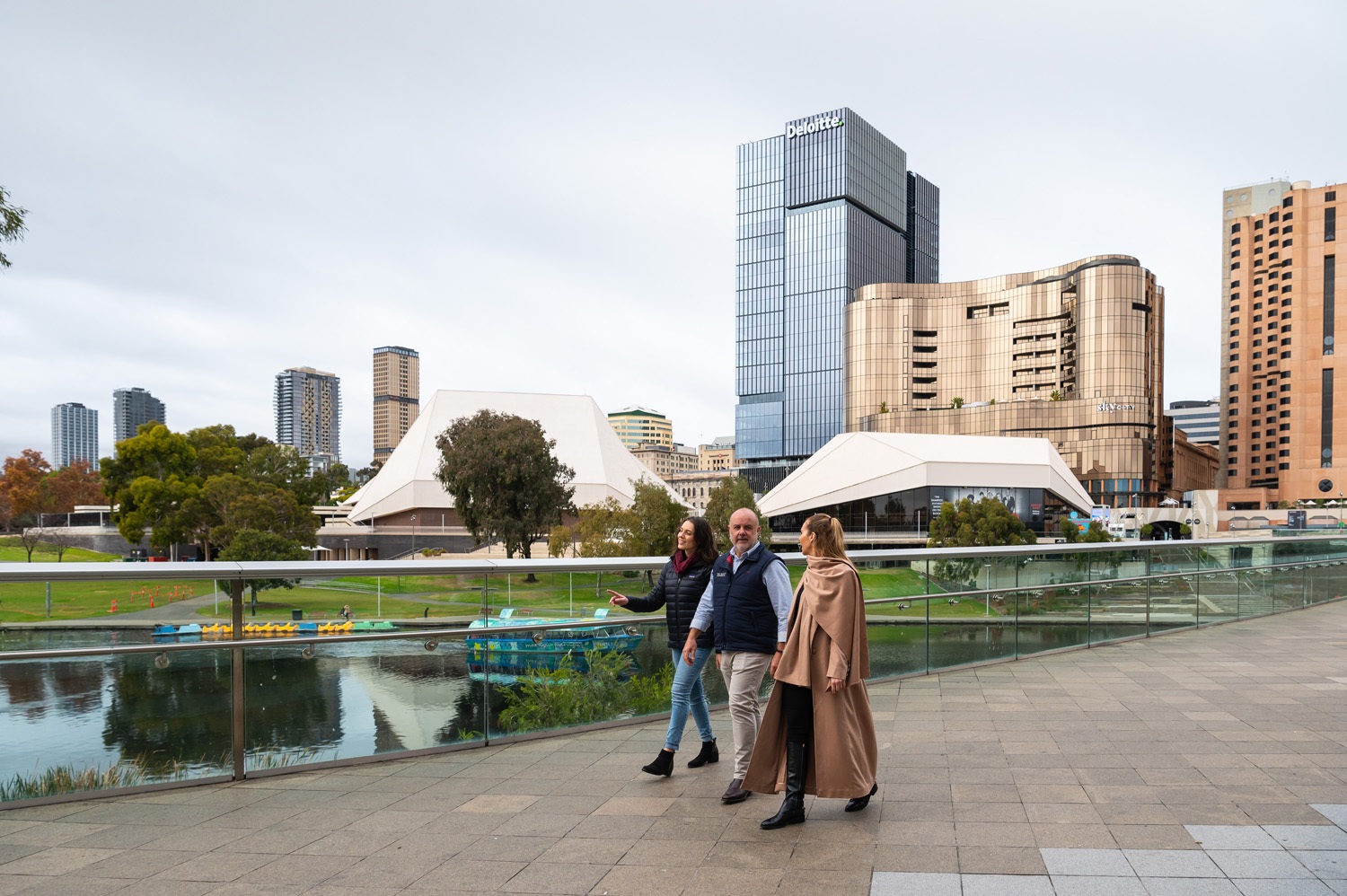 3 people walking along a river in the middle of a city with city building in the background
